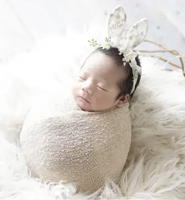 ふわふわの布に包まれて眠るうさぎの耳飾りの新生児