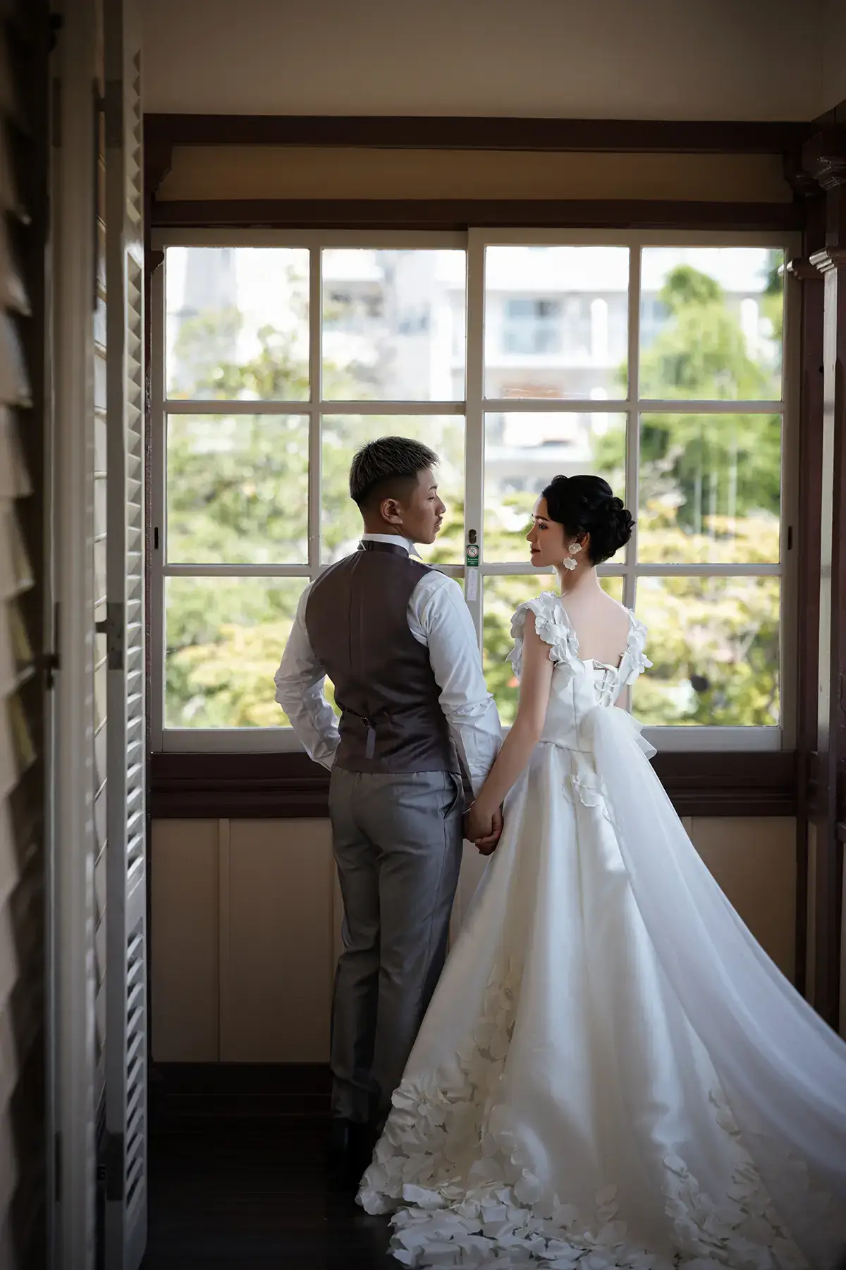 白いドレスとタキシード姿の新郎新婦が窓辺で手を取り合うフォトウェディング撮影
