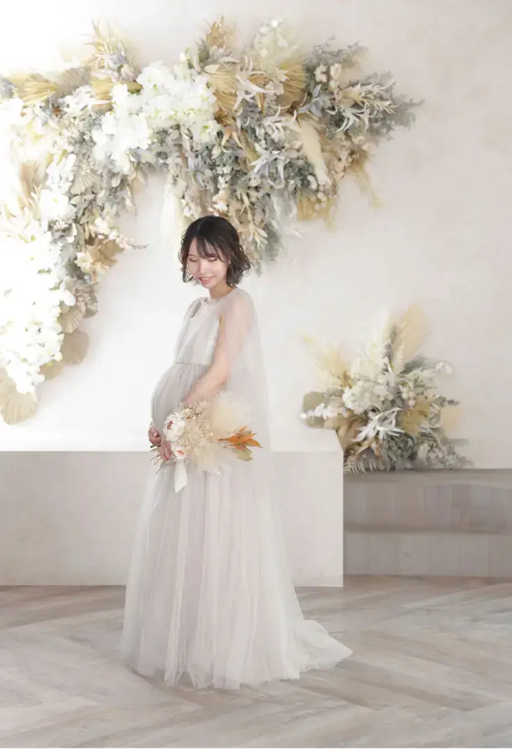 白いフラワーウォールを背景にマタニティドレスで微笑む妊婦さんの記念写真