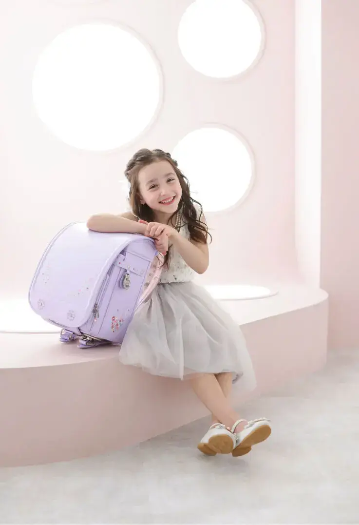 ランドセルに寄りかかって笑顔を見せる女の子と、淡いピンクの背景が可愛らしい入学写真