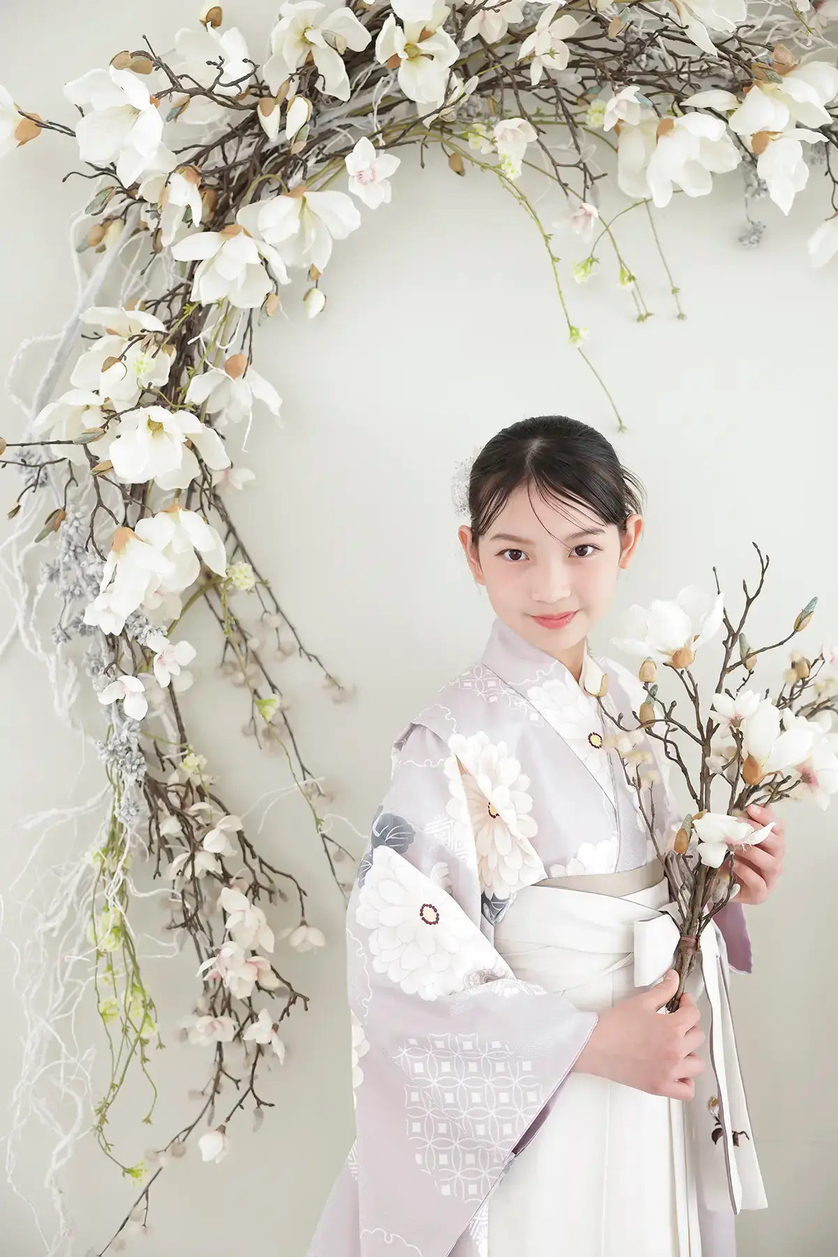 淡い藤色の着物と白い袴で花を手に持つ女の子の小学校卒業記念写真