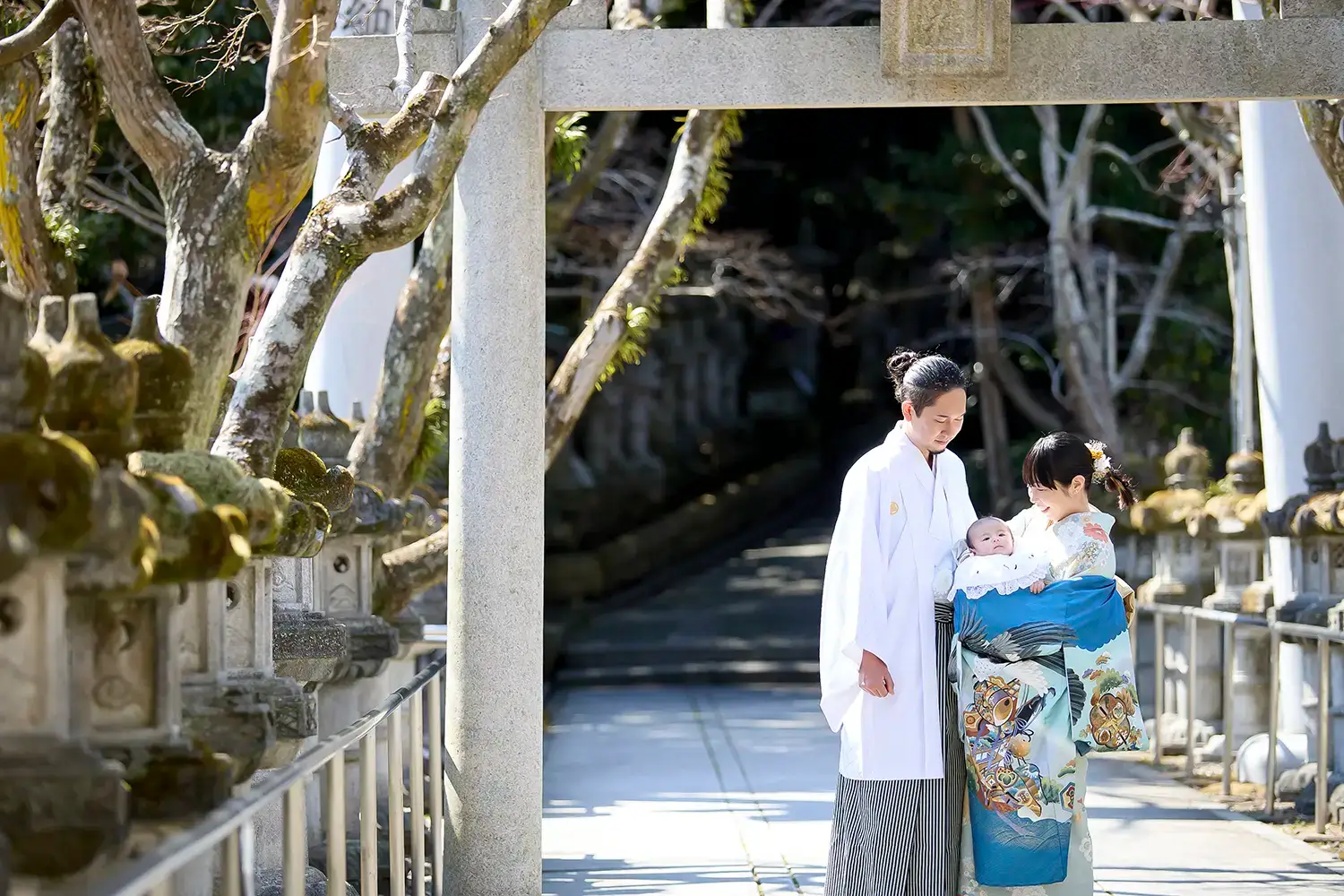 神社の鳥居の下で祝い着をまとった赤ちゃんを囲む家族の温かなお宮参り写真
