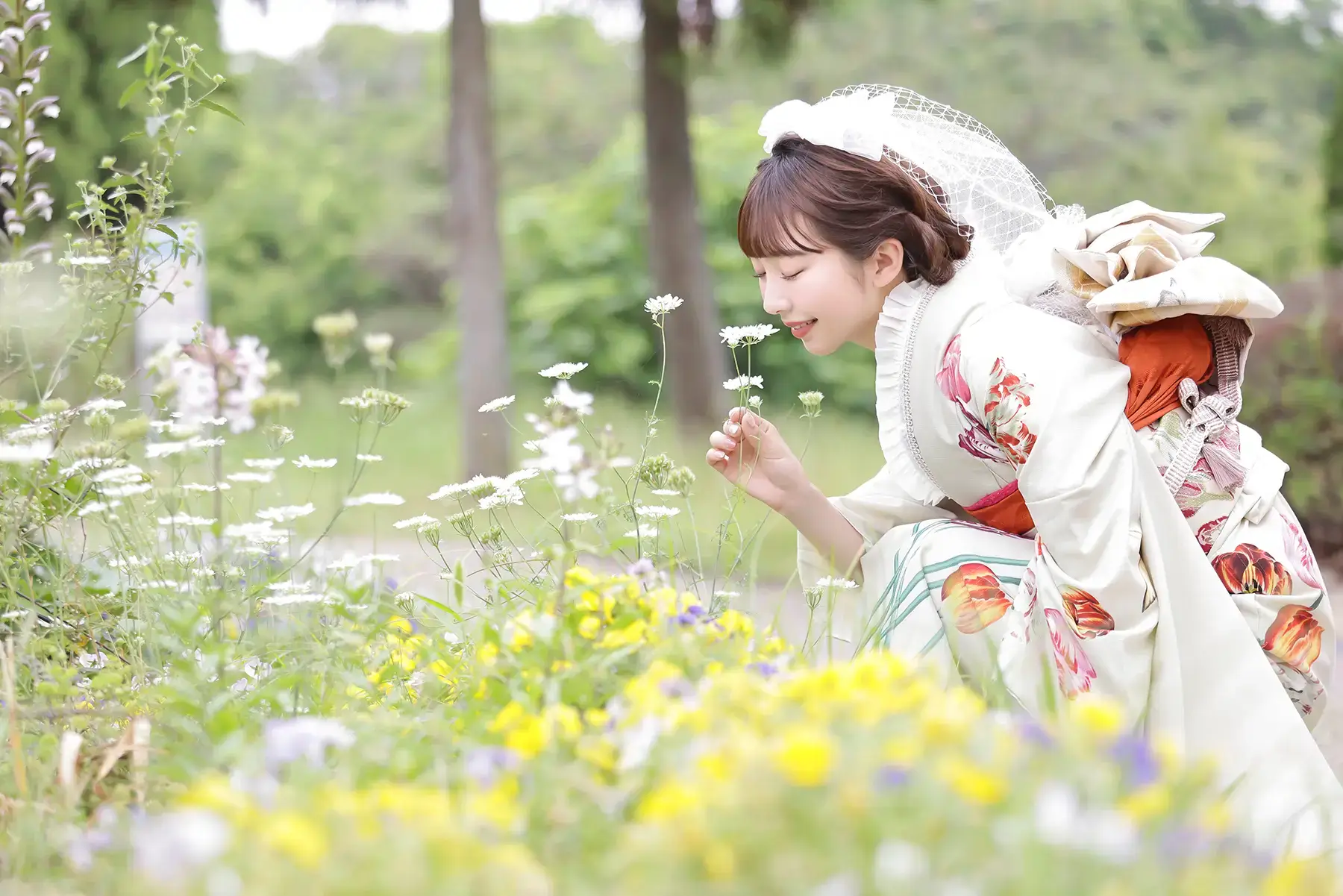 野花に囲まれた振袖姿の女性が笑顔で花に顔を近づける